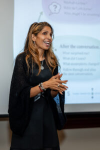 A smiling woman with long highlighted hair, wearing a black dress and dark knit shawl with silver jewelry, gestures mid-presentation in front of a projector screen showing a slide titled 